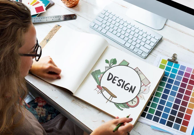 A woman with sitting at a desk with computer keyboard and color palette sheet visible, looking at a notebook with a graphic centered around the word & "design".