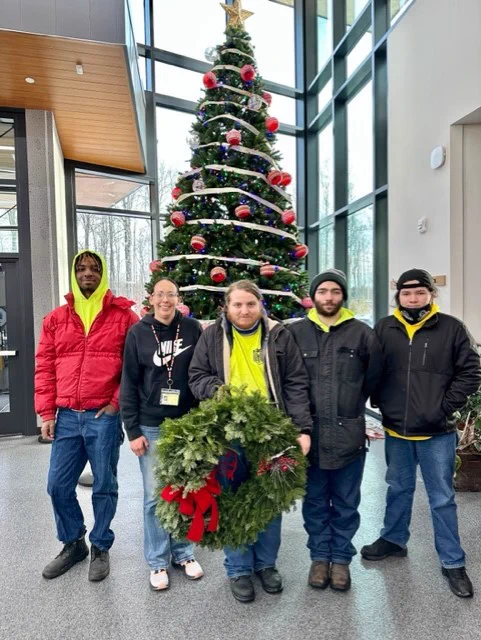 A group of five people pose in front of a Christmas tree. The person in the middle is holding a wreath. 

