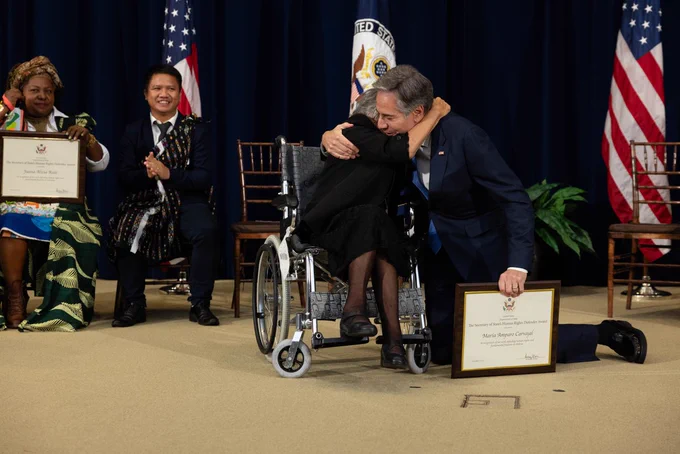 Secretary Blinken kneels down and hugs a woman who is seated in a wheelchair as two people are seated and clapping in the background.
