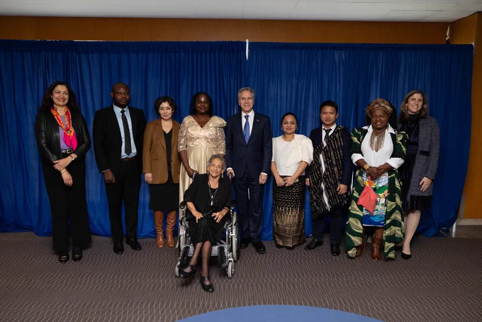 Secretary Blinken stands at the center of a group of people as they pose for a photo in front of a blue curtain.