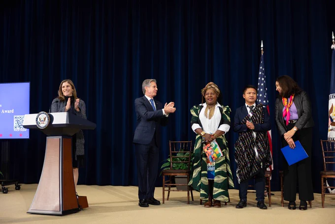 Secretary Blinken claps as he faces a group of people standing on a stage receiving an award for their human rights work. A woman also claps while standing behind him at a lectern.