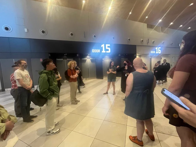 A group of volunteer stands in a modern lobby near doors labeled 15 and 16, attentively listening to an orientation or induction session led by the volunteers coordinator. The setting conveys preparation and teamwork.