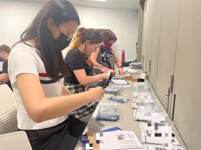Three volunteers are assembling name tags into plastic bags at a neatly organised table, focused and working collaboratively.