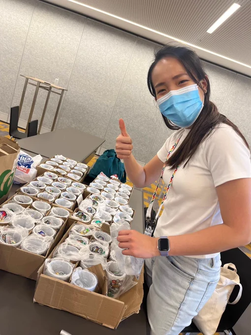 A volunteer wearing a face mask and a white T-shirt gives a thumbs-up while standing next to a table filled with neatly arranged PyCon cups containing various items. The scene captures a cheerful and productive atmosphere.
