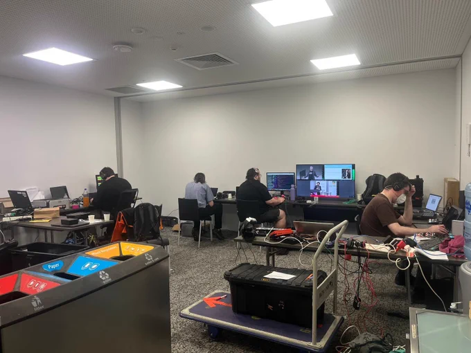 Volunteers from the AV team are working behind the scenes in a room equipped with multiple monitors, laptops, and technical equipment. The team members are focused on their tasks, ensuring smooth operations, while the setup reflects a highly organised and professional environment.