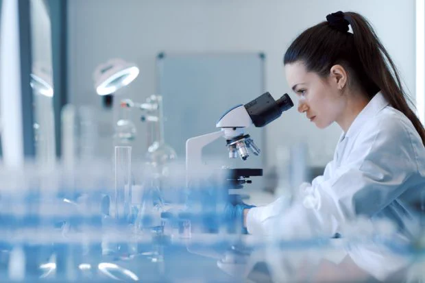 Scientist using a microscope in a laboratory setting, surrounded by various laboratory equipment.