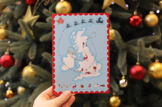 A person holds the GCHQ Christmas Challenge card with a map of the United Kingdom in front of a Christmas tree. The calendar features festive symbols and text in a playful font.