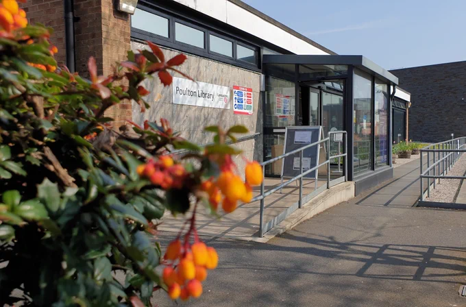 An image of Poulton library on a sunny day. On the far left of the image is a large plant with green leaves and orange petals. You can then see the building of Poulton library filling the rest of the image. There is a ramp leading into the building with grey railings to the right of it and the buildings  windows and doors have black frames.