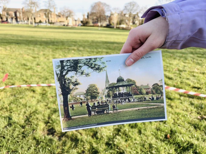 Photo of a girl’s hand holding a historic coloured photo showing Victoria Park in the early 1900s  with people gathered around a bandstand. The photo was taken in the park, grass in the background and red and white hazards tape marking out the location of the bandstand.