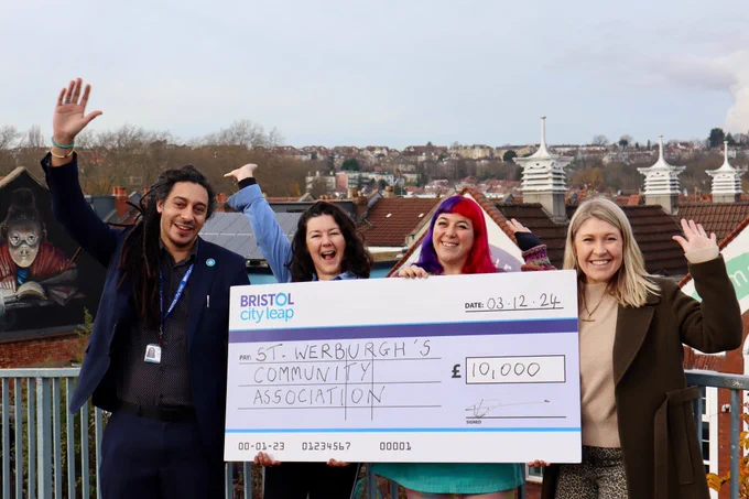 Four individuals standing outside St Werburghs Community Centre, proudly holding a large cheque for £10,000, presented to the centre from the Bristol City Leap Community Energy Fund. The photo captures their smiles and the community spirit.