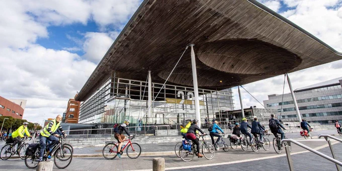 Group of cyclists riding past the Senedd, also known as the Welsh Parliament building, in Cardiff. The building features a distinctive sloping wooden roof.