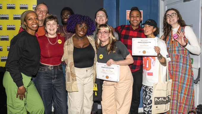 Group of 10 people of mixed ethnicities and genders standing close together smiling. 2 are holding certificates. A multitrack branded screen in yellow can be seen on the left.