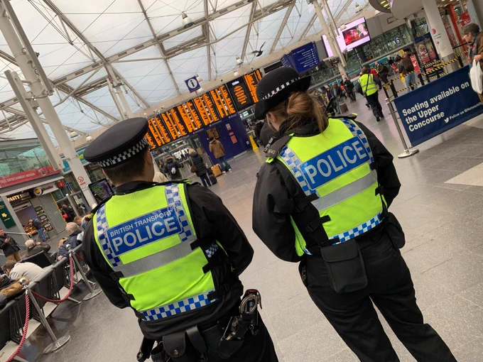 Two BTP officers in full uniform face an information board in a busy train station