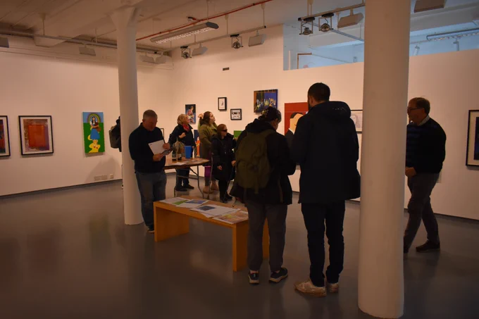 Groups of people gather in a gallery with white walls and colourful artworks on the walls. There is a table in the far back with refreshments and cups on it and a bench in the middle with some paper sheets.