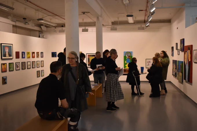 People talking to each other in a gallery space with white walls and colourful artworks. The left wall has a series of square vinyl sized artworks. The other walls have a mix of small and big pieces.