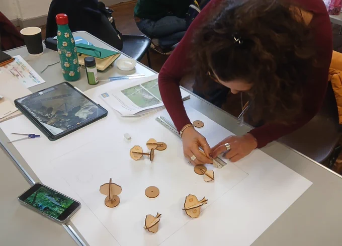 A woman measures a piece of paper that has 3D printed trees and a reference photo of a garden laying on it. 