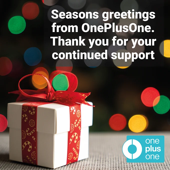 Image shows a white gift box tied with a red ribbon featuring candy cans and sweets printed in gold. The background is dark, with a blurred, multi-coloured dotty pattern, resembling Christmas lights. The OnePlusOne logo is in the bottom right-hand corner.