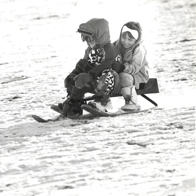 A black and white photo of two children in snowsuits on a sled. The child in front is wearing a patterned snowsuit with a hood, plus goggles. The child behind has long hair and a light colored headband. 