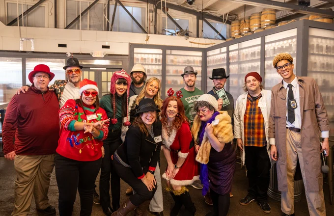Group of people in festive attire posing happily at a party, some wearing Christmas-themed outfits and others in unique costumes, in an indoor setting.