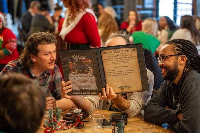 Three people sit at a table, smiling, while one holds a book titled "How to Play a Suspect". They are in a lively, festive venue with other people visible in the background.