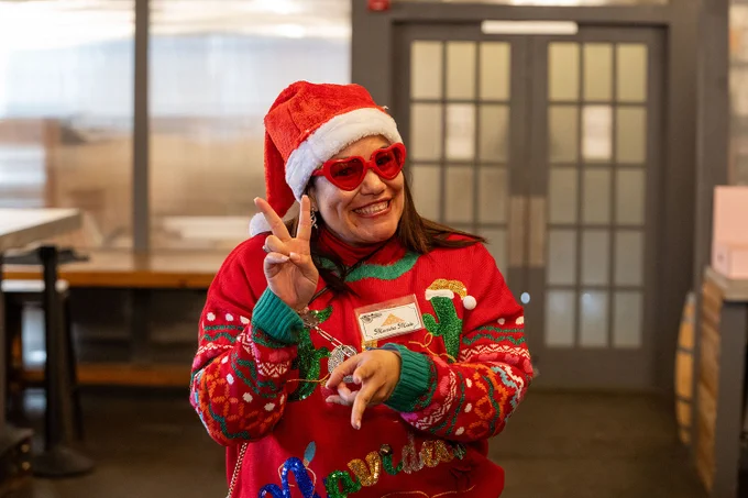 Person wearing a festive sweater and sunglasses, a Santa hat, and handcuffs gives a peace sign while smiling.