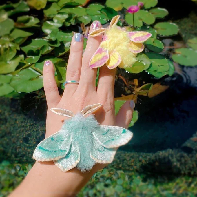 The image shows a hand holding two handmade moth brooches. 

Moth 1

Moth 2

Color

Pink and yellow

Turquoise and silver

Position

On the index finger

On the palm

