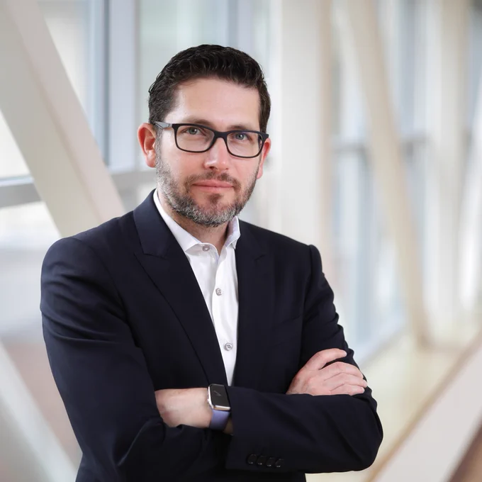 Professional portrait of Dr. Alejandro Quiroga in a business suit, standing with crossed arms in a modern office setting.