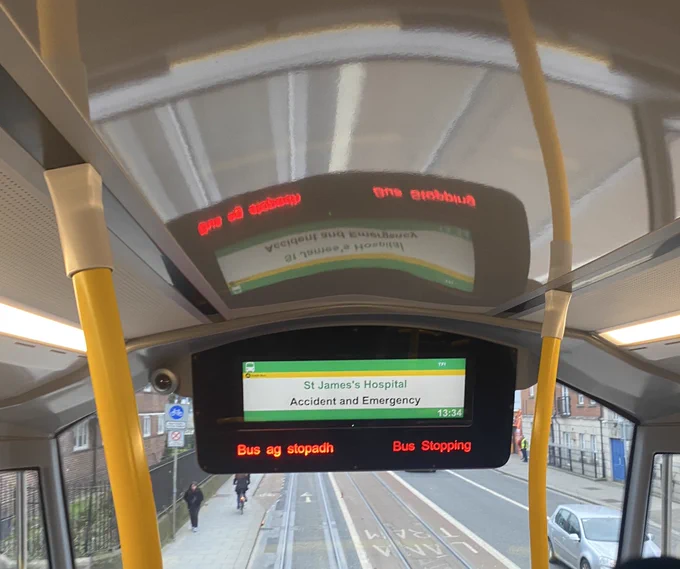 Display screen on upper deck of a bus indicating next stop: St James’s Hospital. View of road ahead below framed by yellow vertical handrails of the bus.