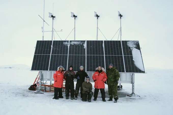 Six personnes, deux portant un manteau d’hiver orange, quatre portant des vêtements d’hiver plus foncés, debout devant un grand panneau solaire auquel sont attachées des éoliennes. Au-delà du panneau se trouvent un terrain enneigé et un ciel blanc solide.