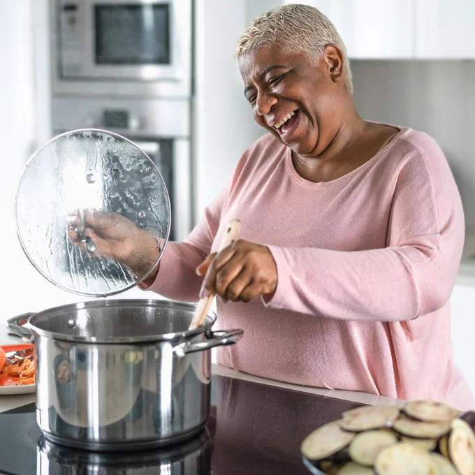 Mature black woman wearing a pink sweater smiles wide as she stirs a pot on an induction stovetop.