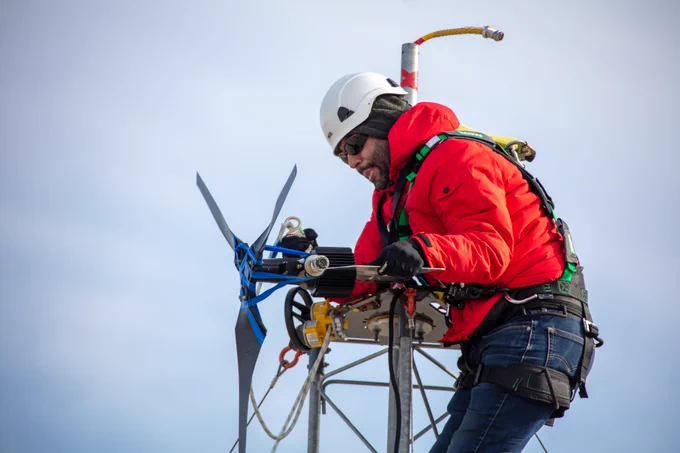 Person in white helmet, orange winter jacket, sunglasses, a harness and blue jeans making repairs at the top of a wind turbine. Solid light blue sky as background.