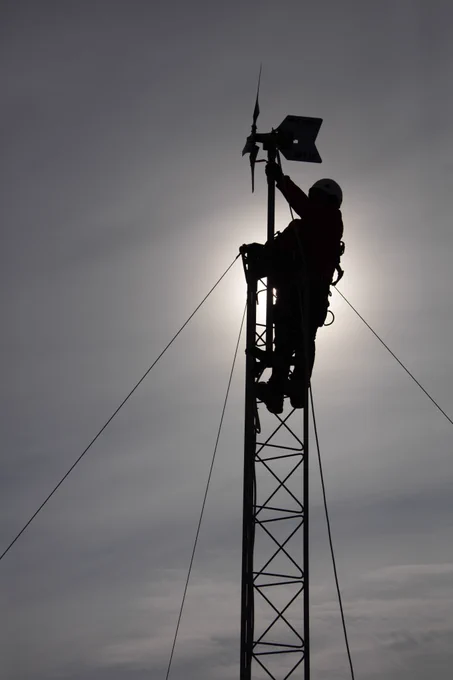 Silhouette of a person at the top of a wind turbine. Blue/grey sky in background.