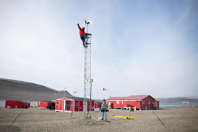Person in orange winter jacket with one hand held upwards, at the top of a wind turbine. At the bottom of the turbine is grassy land and red buildings with white trim. Another individual stands at the bottom of the turbine looking upward. There is a red truck to the left, blue sky in background.