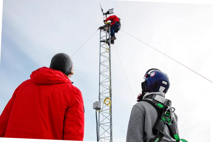 Person in a white helmet and orange winter jacket at the top of a wind turbine. Two other individuals are at the bottom of wind turbine, one in helmet watching while the other supports a line connected to the turbine.
