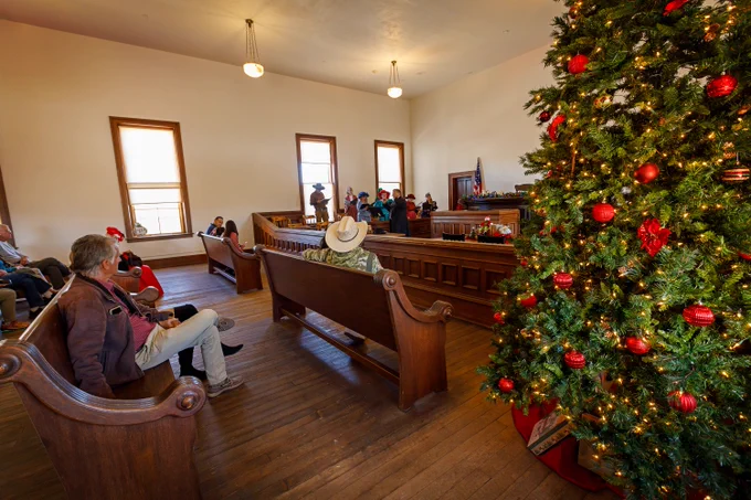 Interior of a historical courtroom decorated for Christmas, featuring a large decorated tree on the right, visitors sitting on wooden benches, and a person standing at the judge's bench in front of a small group.