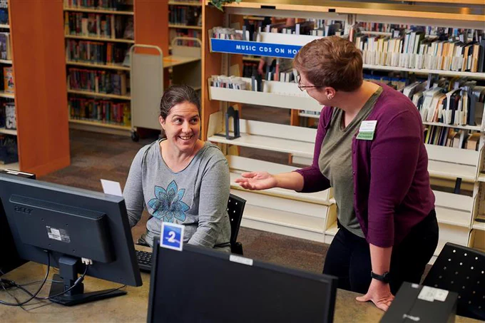 Photo of two people inside a library, one sitting at a computer and smiling and the other standing and facing her.