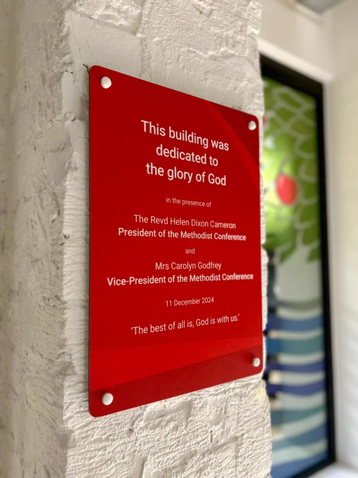 Photo of a red plaque with white text which reads 'this building was dedicated to the glory of God in the presence of The Revd Helen Dixon Cameron, President of the Methodist Conference and Mrs Carolyn Godfrey, Vice - President of the Methodist Conference 11 December 2024. 'The best of all is, God is with us''. Mounted on a white wall.