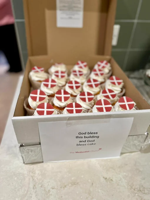 Box of cupcakes decorated with a sugar paper Methodist logo. Taped to the white box containing the cakes is a sign which reads 'God bless this building and God bless cake'. 