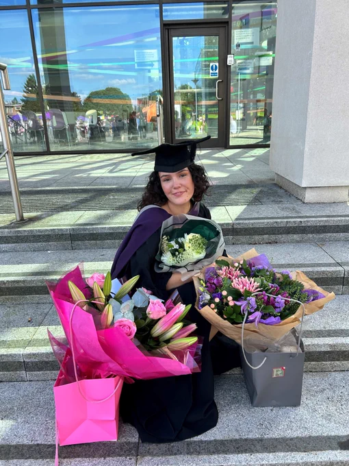 Recent Media and Communications Graduate Lauryn Chandler-Johnson on her graduation day, wearing her cap and gown surrounded by bouquet of flowers as she sits on a step. 