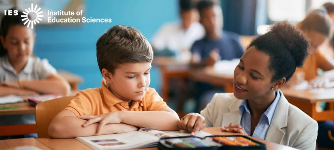An elementary school teacher crouches down next to a student sitting at a desk in a classroom while they read from an open book together.