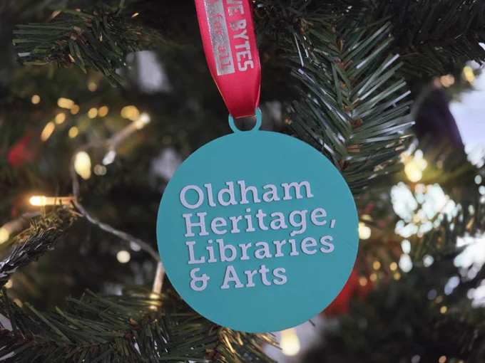 A christmas decoration featuring the logo of Heritage Libraries & Arts hanging on a decorated tree.
