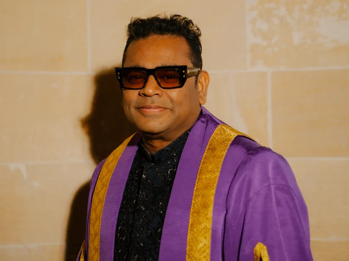A. R. Rahman is smiling toward the camera. He is wearing dark tinted glasses and a purple robe with gold detailing.