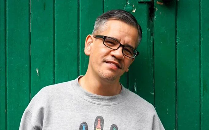 An Asian man with short and grey hair, on a green panelled backdrop, wearing glasses and a grey sweatshirt.