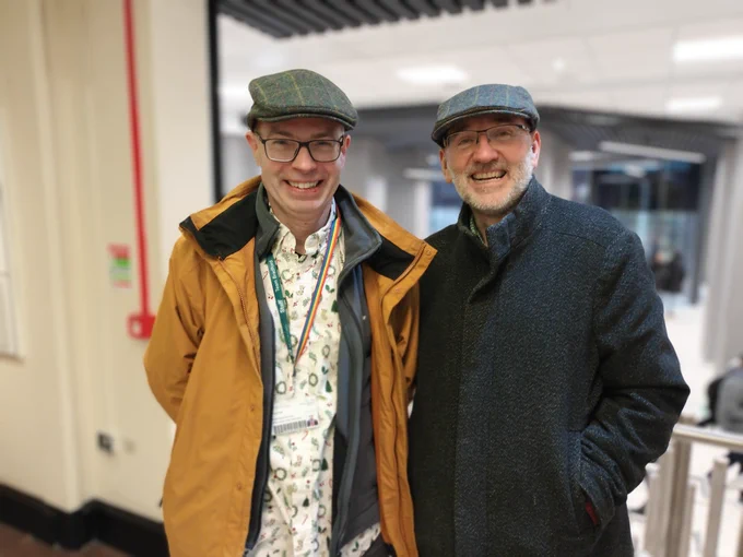 2 bald, bespectacled, middle-aged men in identical flat caps. 
