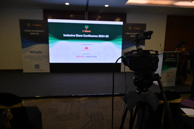 A camera on a tripod is positioned in the foreground, pointed towards a stage with a large screen displaying "Inclusive Stem Confluence 2024-25" under the banner of "I-Stem."