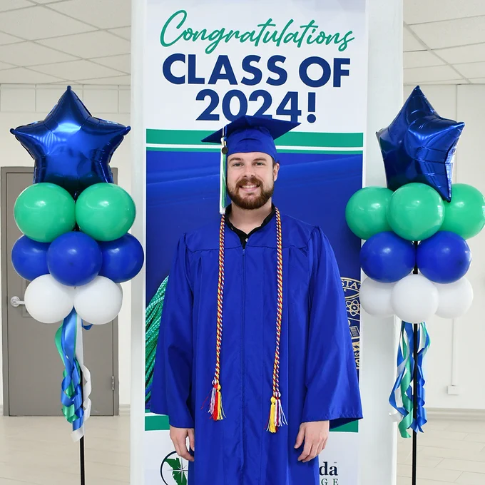 male graduate in blue cap and gown in front of Congratulations Class of 2024! banner with green, blue and white balloons