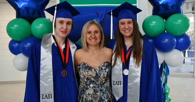 two graduates in blue caps and gowns with women in dress in front of balloons