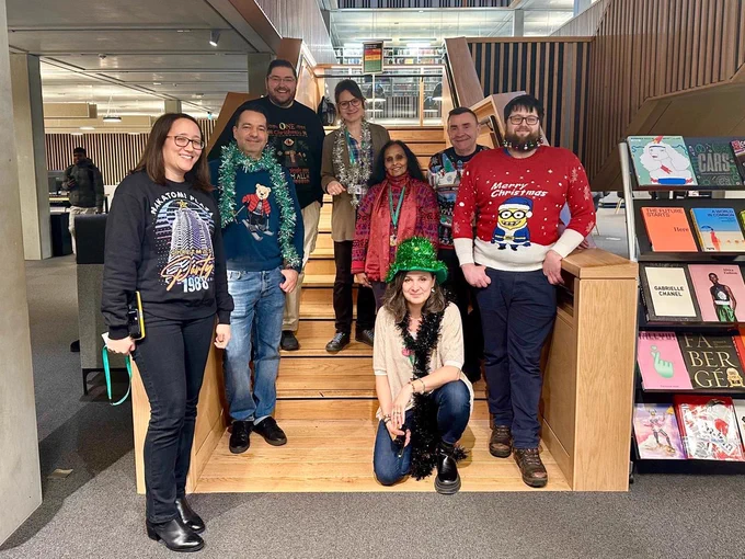 Image shows a group shot of library staff with Christmas jumpers standing on the library staircase 