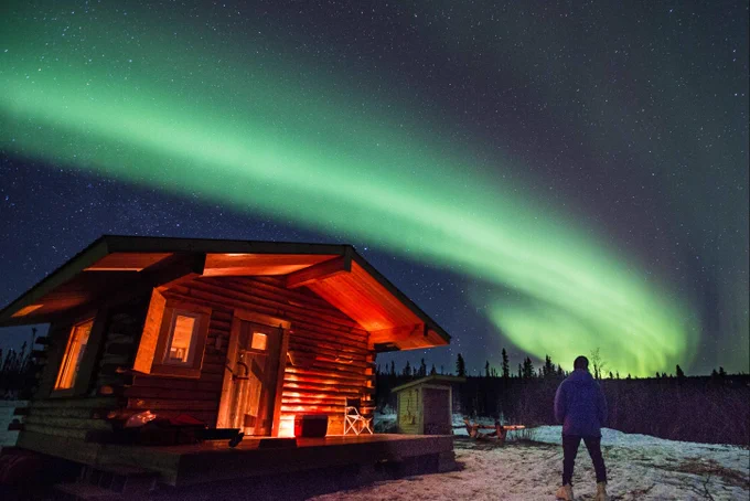 A person stands next to a cabin illuminated in red by light with green lights in the sky overhead