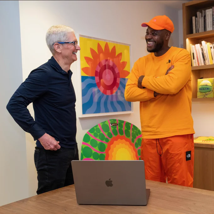 Tim and Yinka stand side by side, facing each other and chatting. Behind them is a tall bookshelf and white wall with some of Yinka’s art displayed. MacBook sits on the table in front of them.